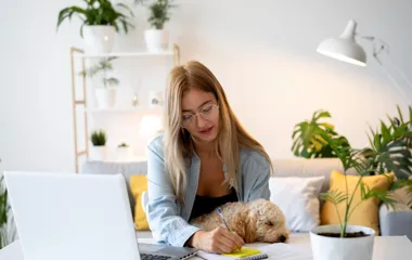 Kobieta w okularach pisze przy laptopie, a na jej kolanach śpi mały pies.