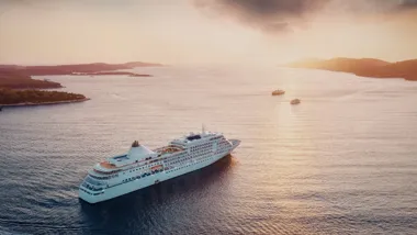 Statek wycieczkowy płynie po morzu o zachodzie słońca, w tle widoczne wyspy.