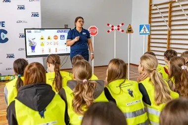 Kobieta w niebieskiej koszulce prowadzi lekcję bezpieczeństwa ruchu drogowego i kolejowego dla dzieci w żółtych kamizelkach odblaskowych w sali gimnastycznej.
