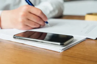 Dłoń osoby piszącej długopisem na dokumentach leżących na drewnianym stole, obok smartfon.