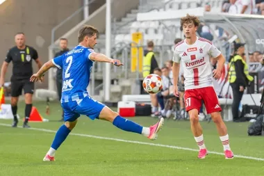 Piłkarz w niebieskim stroju z numerem 2 uderza piłkę podczas meczu ligowego na stadionie, obok niego stoi zawodnik w biało-czerwonym stroju z numerem 37.